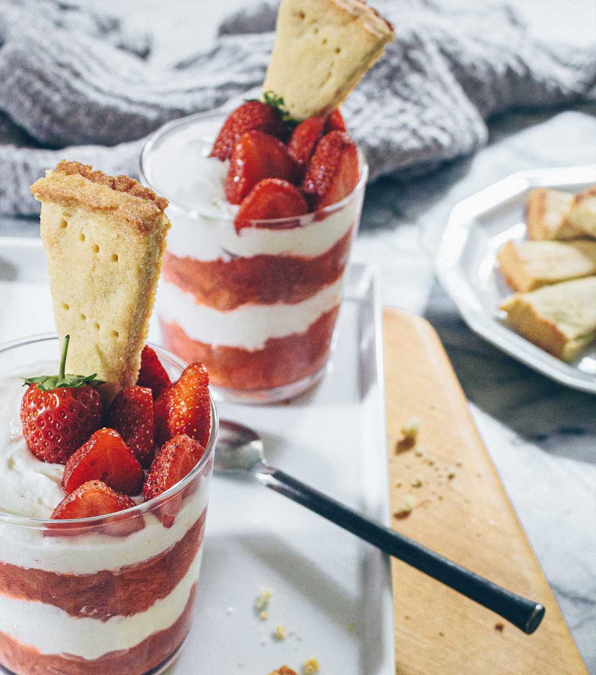 Glass jars filled with Strawberry Rhubarb compotes layered with Cheesecake Cream, with Shortbread Cookie pieces and strawberries sticking in the top.
