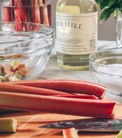 A cutting board with rhubarb stalks being chopped to make rhubarb gin. In the background there is a bottle of Barr Hill Gin and a glass bowl with chopped rhubarb mixed with sugar.