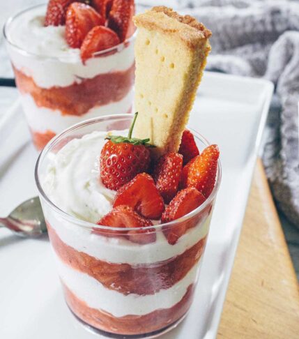 Glasses filled with red and white layers of strawberry rhubarb compote and cheesecake cream, topped with fresh strawberries and with a shortbread stuck in the top.