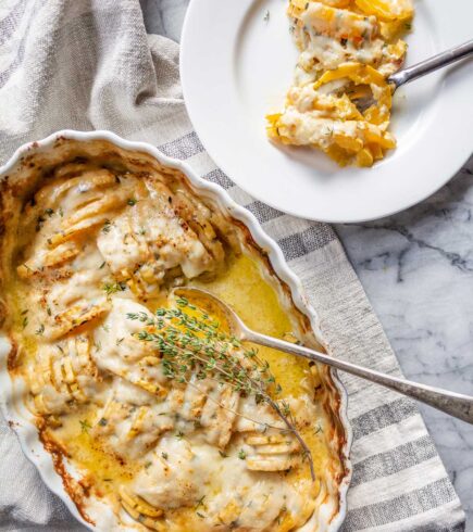 An white oval ceramic serving dish filled with Delicata Squash Gruyere Gratin, covered in melted cheese and fresh thyme. Next to this is a white plate with a serving of the gratin.