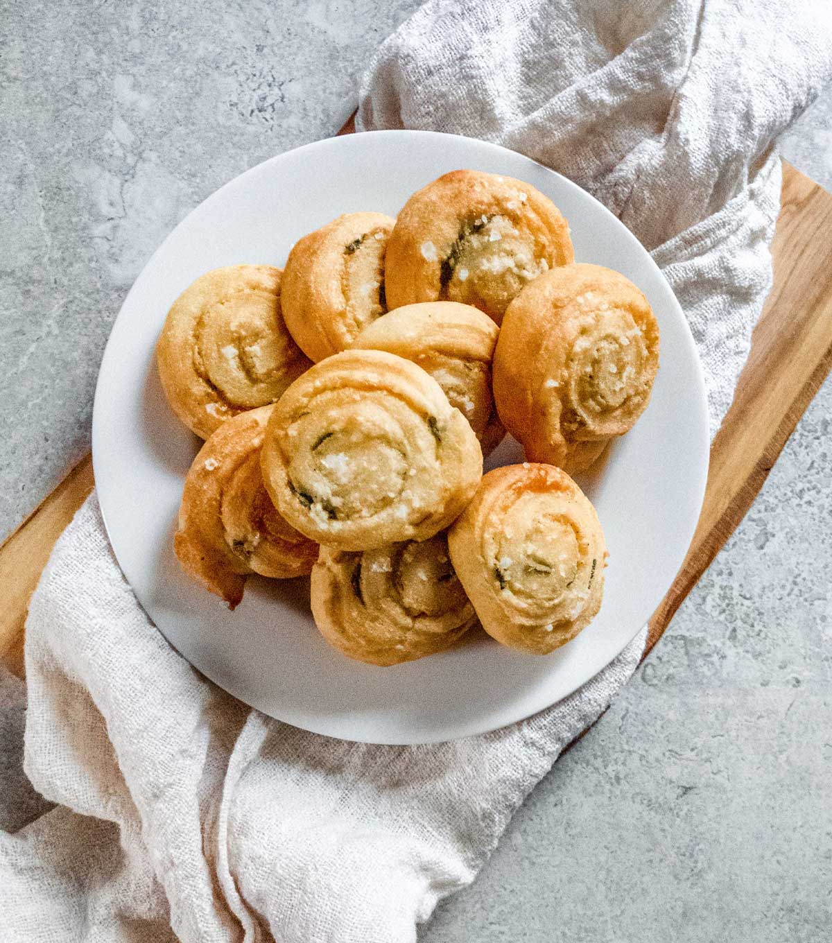 A white plate on top of a linen napkin piled high with Parmesan Sage Pinwheel rolls.