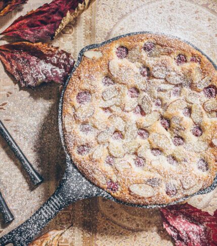 A cast iron skillet filled with a cake studded with raspberries and almonds and sprinkled with confectioner's sugar. Fall leaves are strewn about.