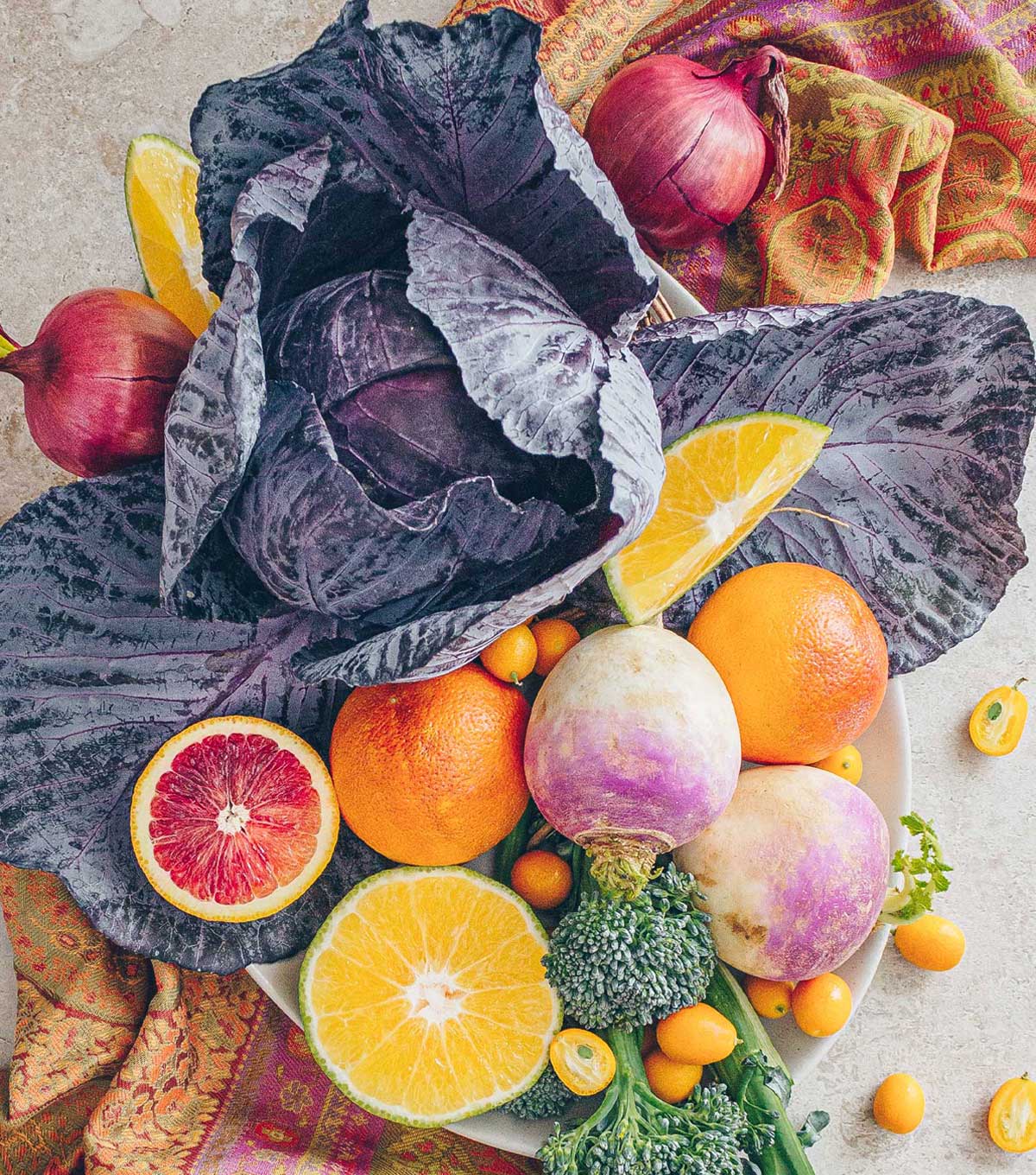 A display of seasonal fruits and vegetables for February: purple cabbage, turnips, broccoli, onions, blood oranges, ugly fruit, and kumquats on a paisley runner.