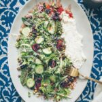 An oval platter on an indigo printed tablecloth filled with arugula and spring greens, cucumber, and cherries with a long smear of whipped feta and drizzled with a red pomegranate balsamic vinaigrette.