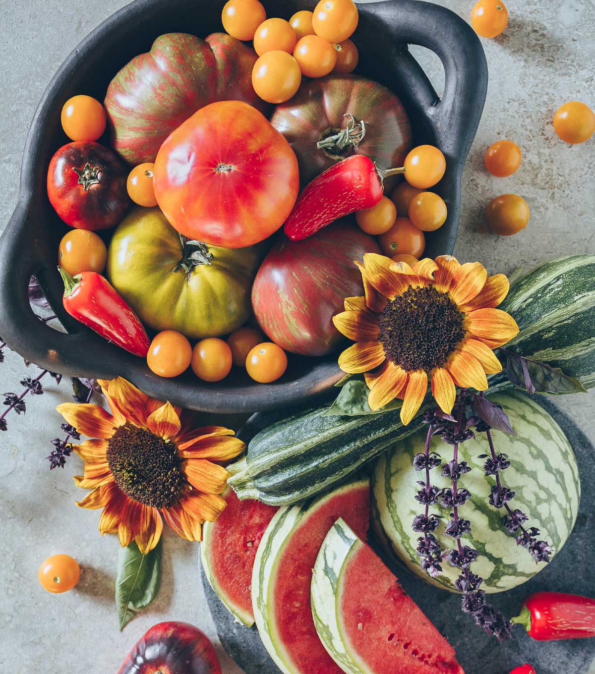 A tabletop display of seasonal eating produce for August. A bowl filled with heirloom tomatoes, zucchini squash, peppers, sunflowers, herbs, and watermelon.