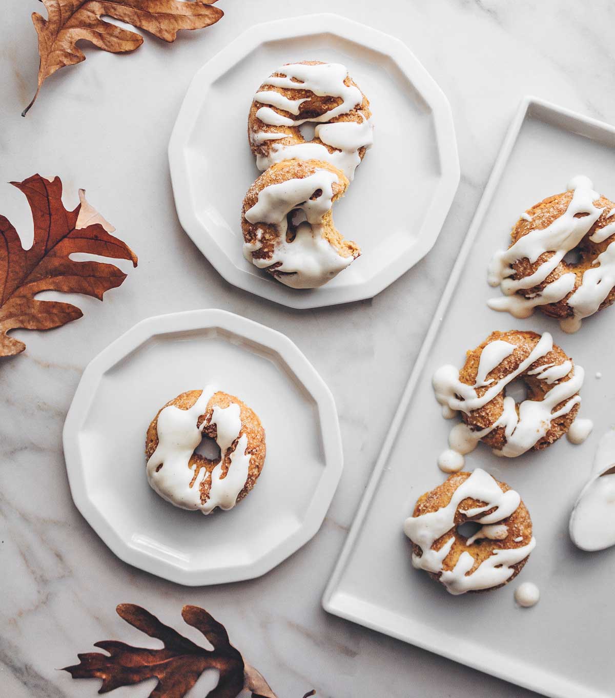 A marble countertop with white plates containing pumpkin spice donuts drizzled with cream cheese glaze. Fall oak leaves are scattered about.