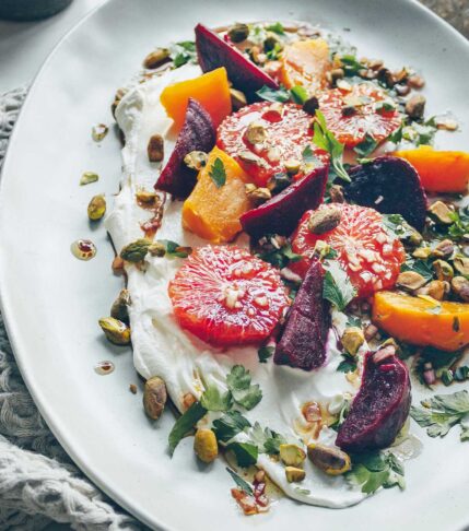 An oval plate with a long smear of Labneh yogurt cheese covered in roasted red and gold beets and slices of blood oranges sprinkled with chopped pistachios and parsley.