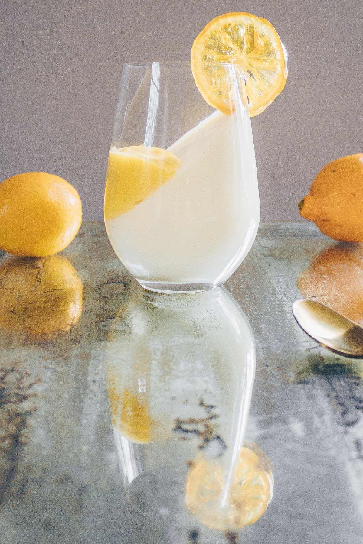 An antiqued mirrored table with a stemless wine glass filled with Yogurt Pannacotta at an angle with a well of Meyer lemon curd on the side and a candied lemon on the rim.