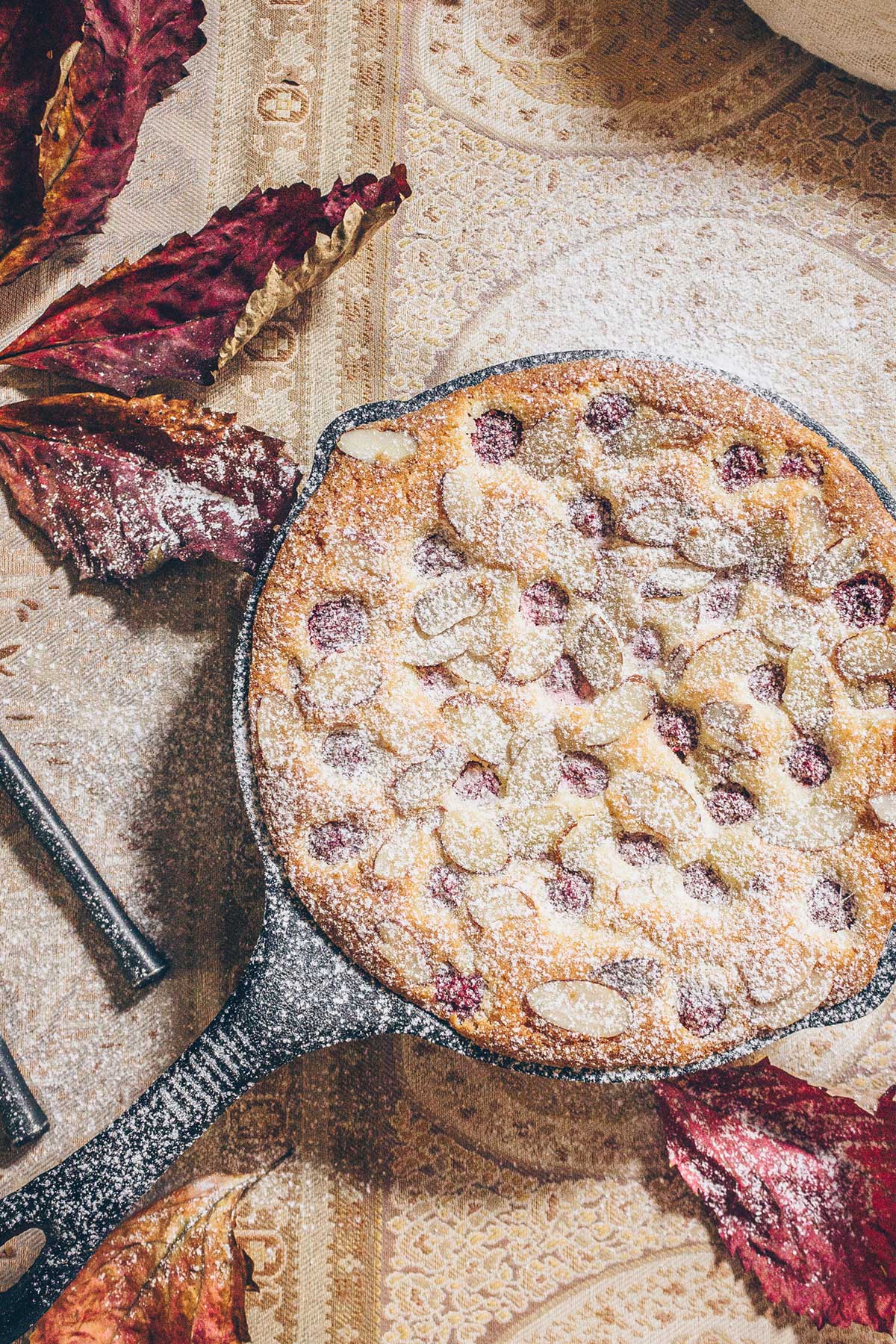 A Raspberry Almond cake baked in a cast iron skillet on a table with dried burgundy red leaves and a pale sand colored paisley tablecloth. The cake is being dusted with powdered sugar, falling from above.