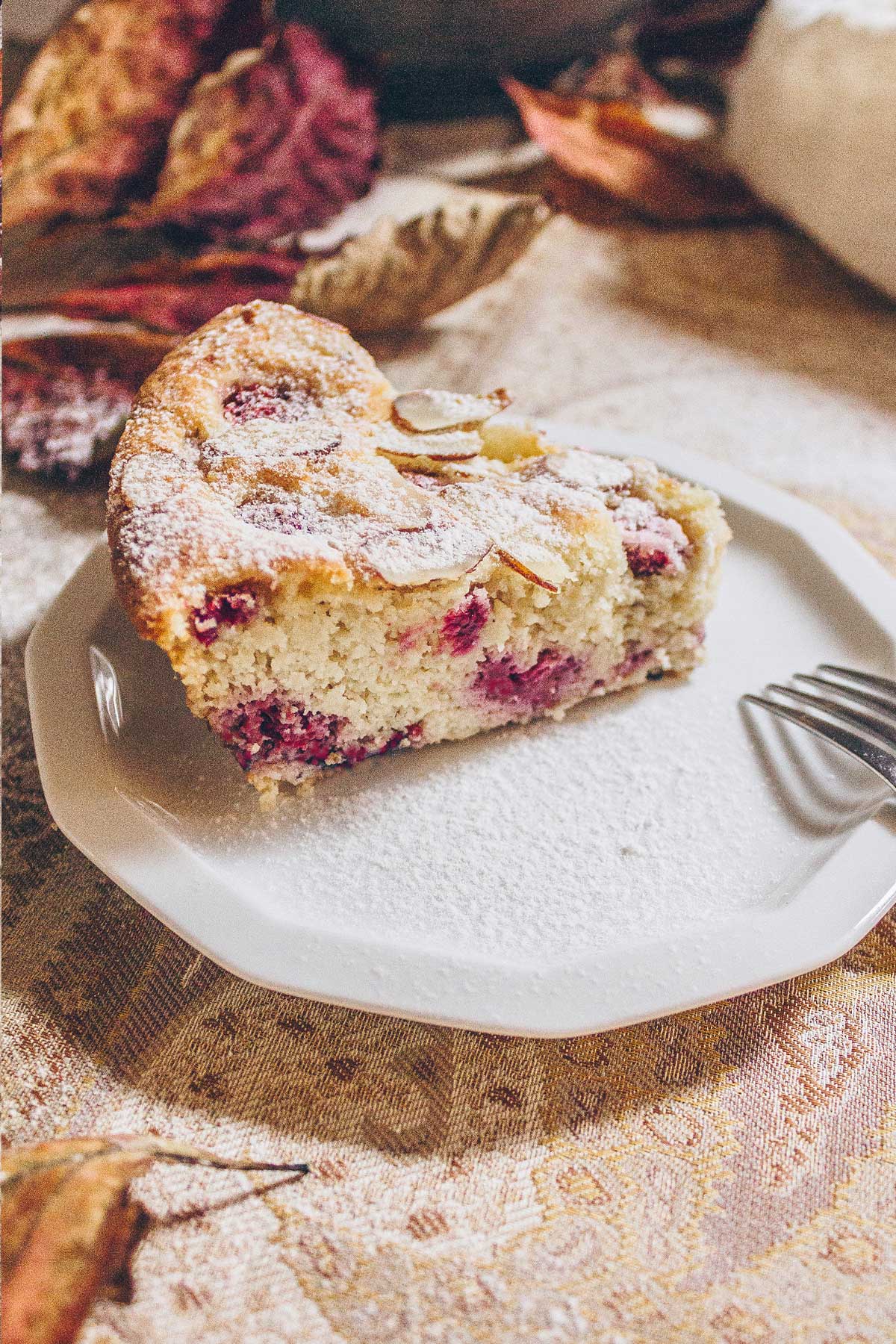 A slice of Raspberry Almond cake on a white plate on a table with dried burgundy red leaves and a pale sand colored paisley tablecloth. The cake is dusted with powdered sugar.