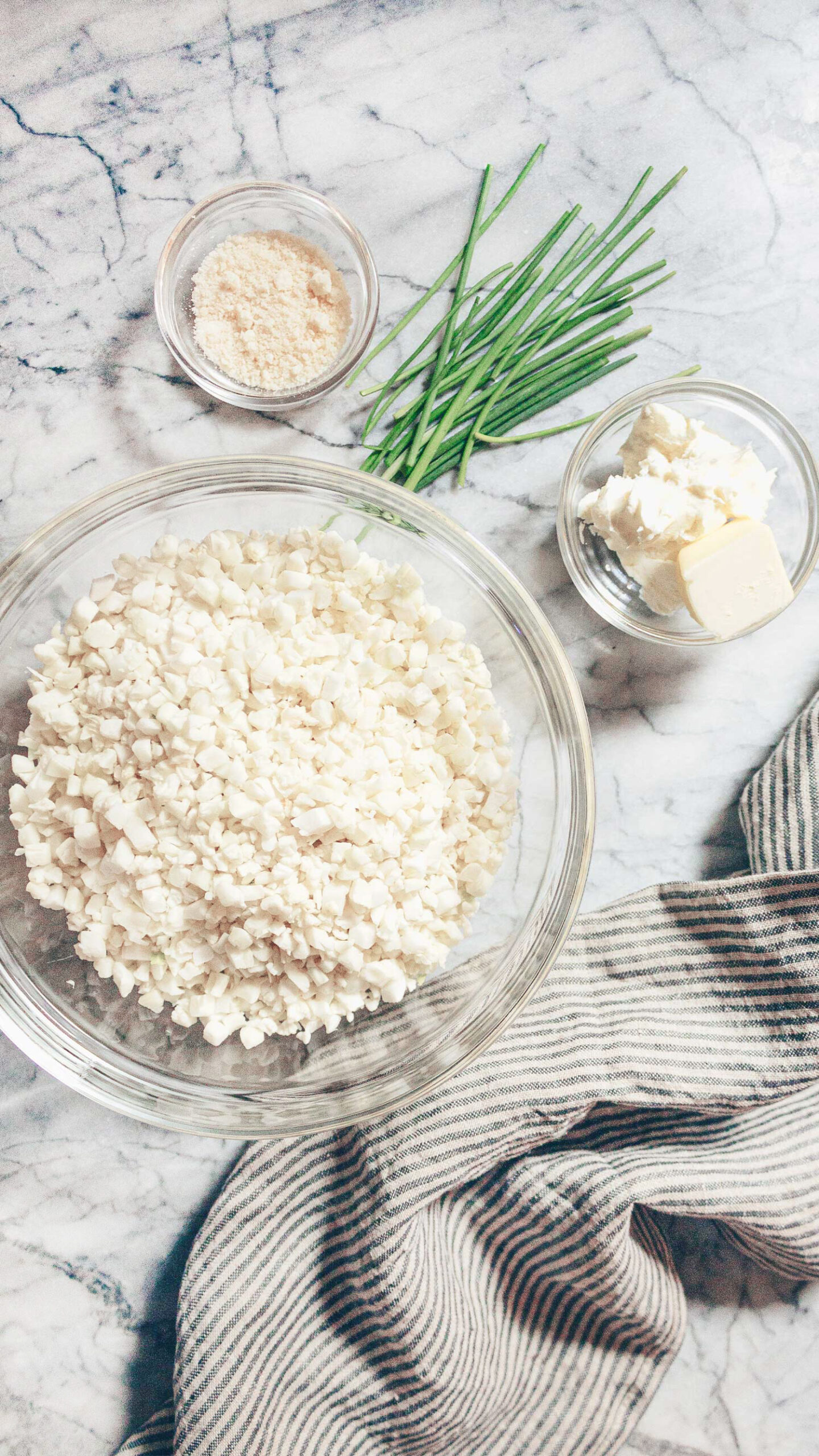 The ingredients for Whipped Cauliflower With Mascarpone displayed in bowls on a marble slab: riced cauliflower, mascarpone, butter, parmesan cheese. 