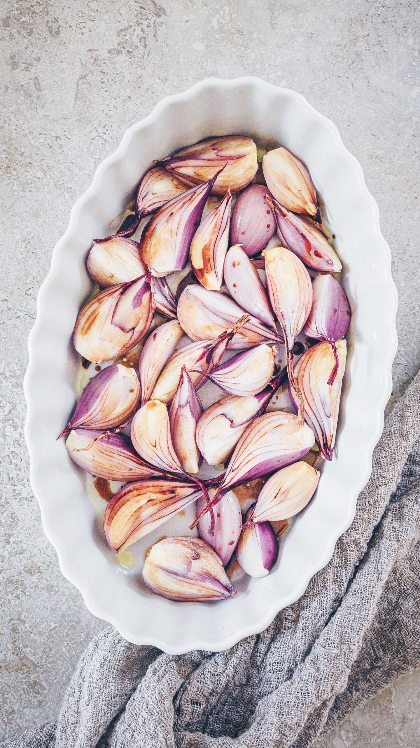 A white porcelain fluted oval baking dish with peeled shallots coated in olive oil and balsamic vinegar.