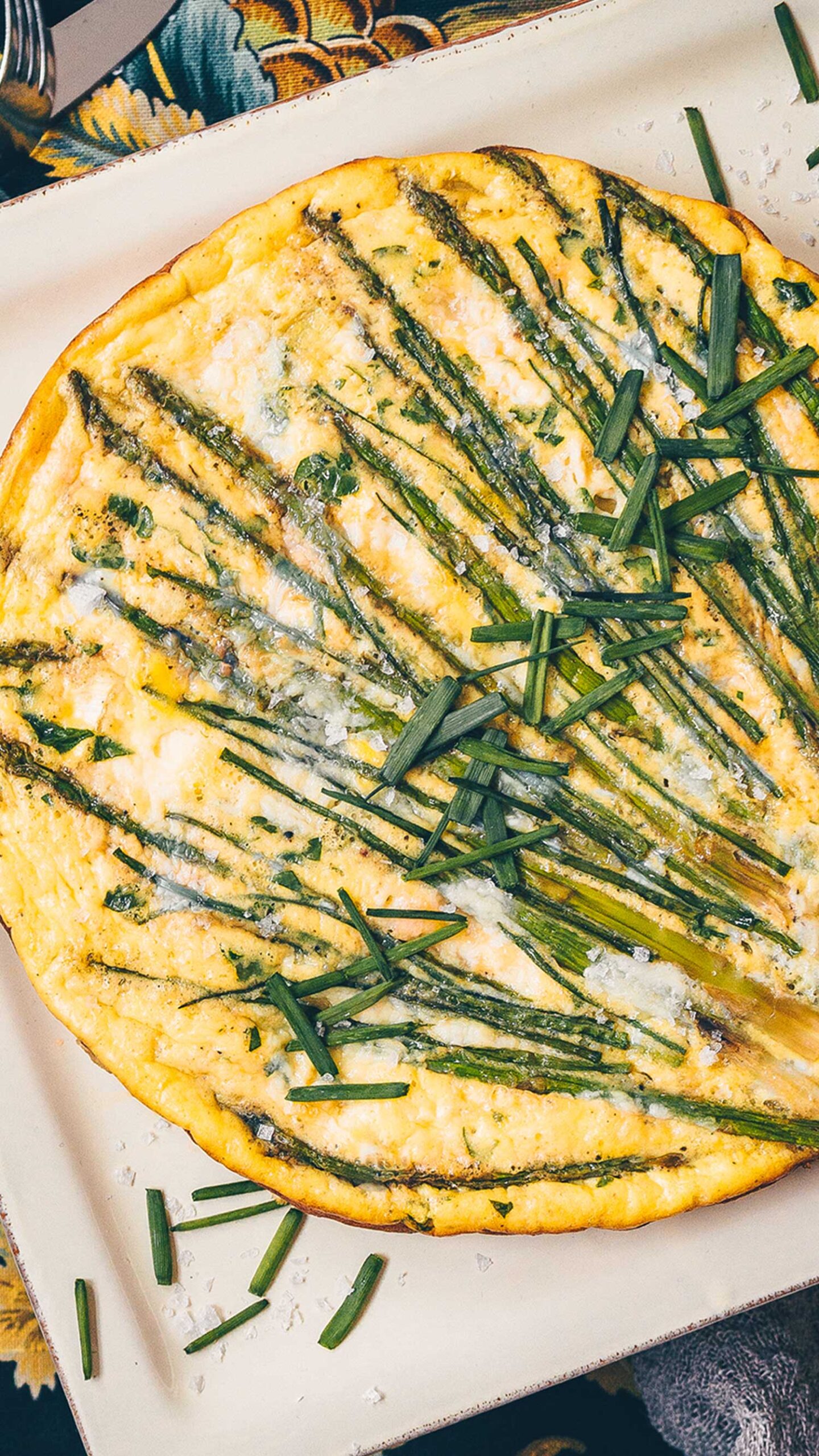 A close up of a large Asparagus frittata with the asparagus spears laid across the top in a wild pattern, and fresh chives across the top.