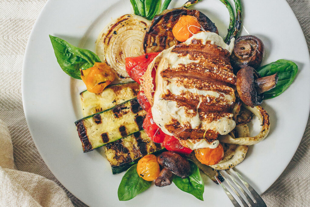 A plate of layered grilled summer vegetables topped with a large piece of grilled smoked mozzarella cheese.