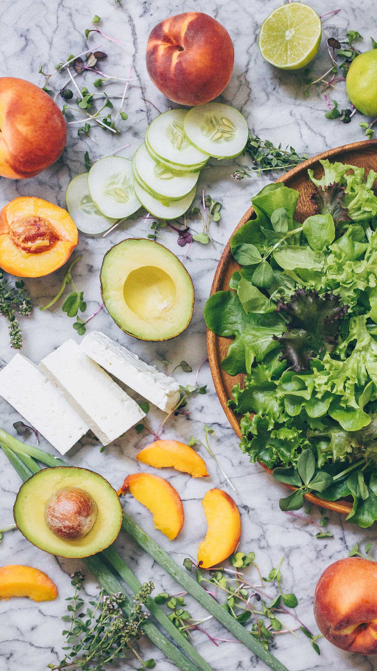 The ingredients for Peach, Avocado and Feta Cheese Salad laid out on a marble counter: peaches, avocado, feta cheese, cucumber, salad greens, limes, and thyme.