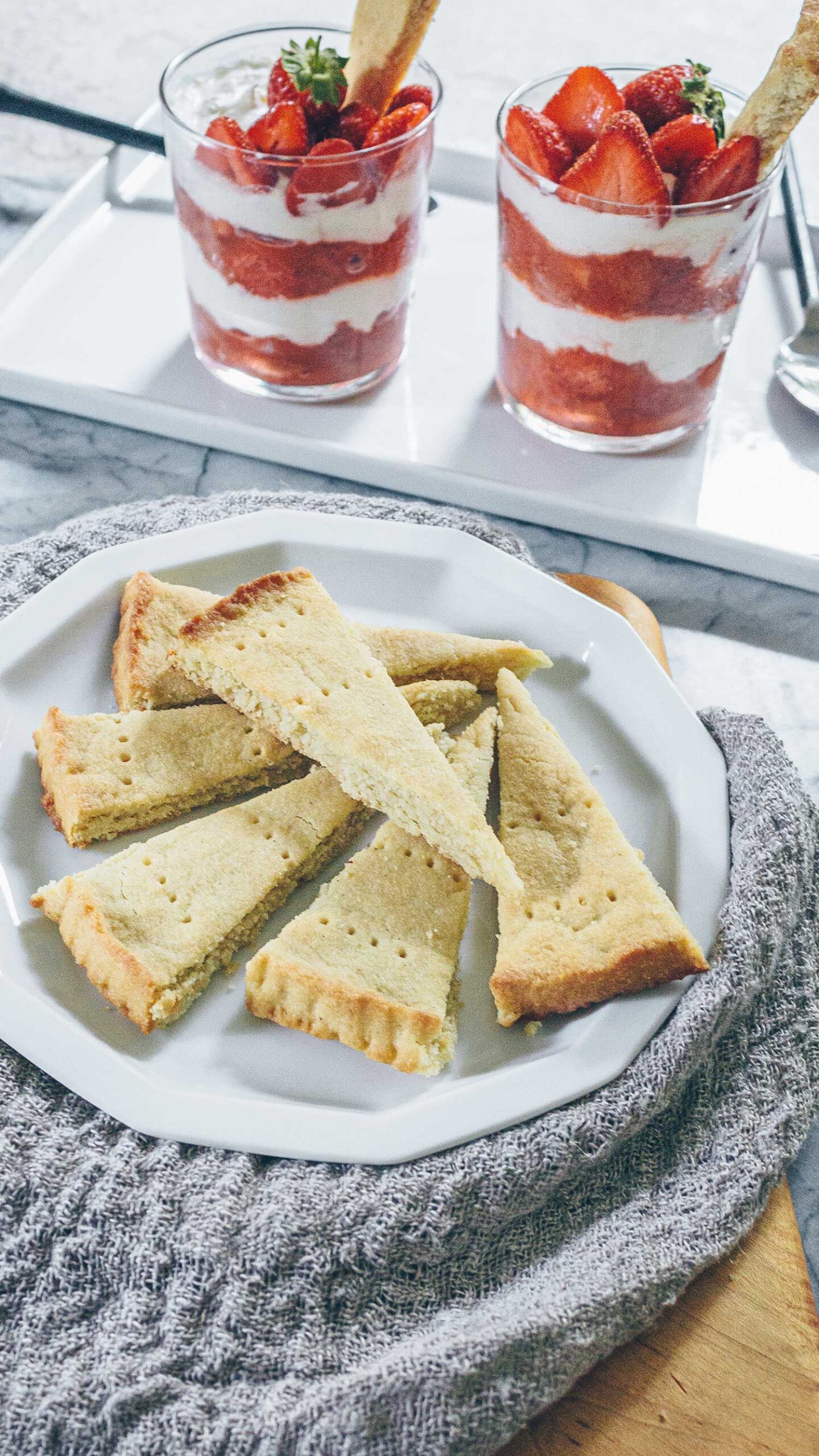 A plate of shortbread cookies next to two clear glasses layered with red Strawberry Rhubarb compote and creamy Cheesecake Cream topped with fresh strawberries and with a shortbread cookie stuck in the top to use as a spoon.