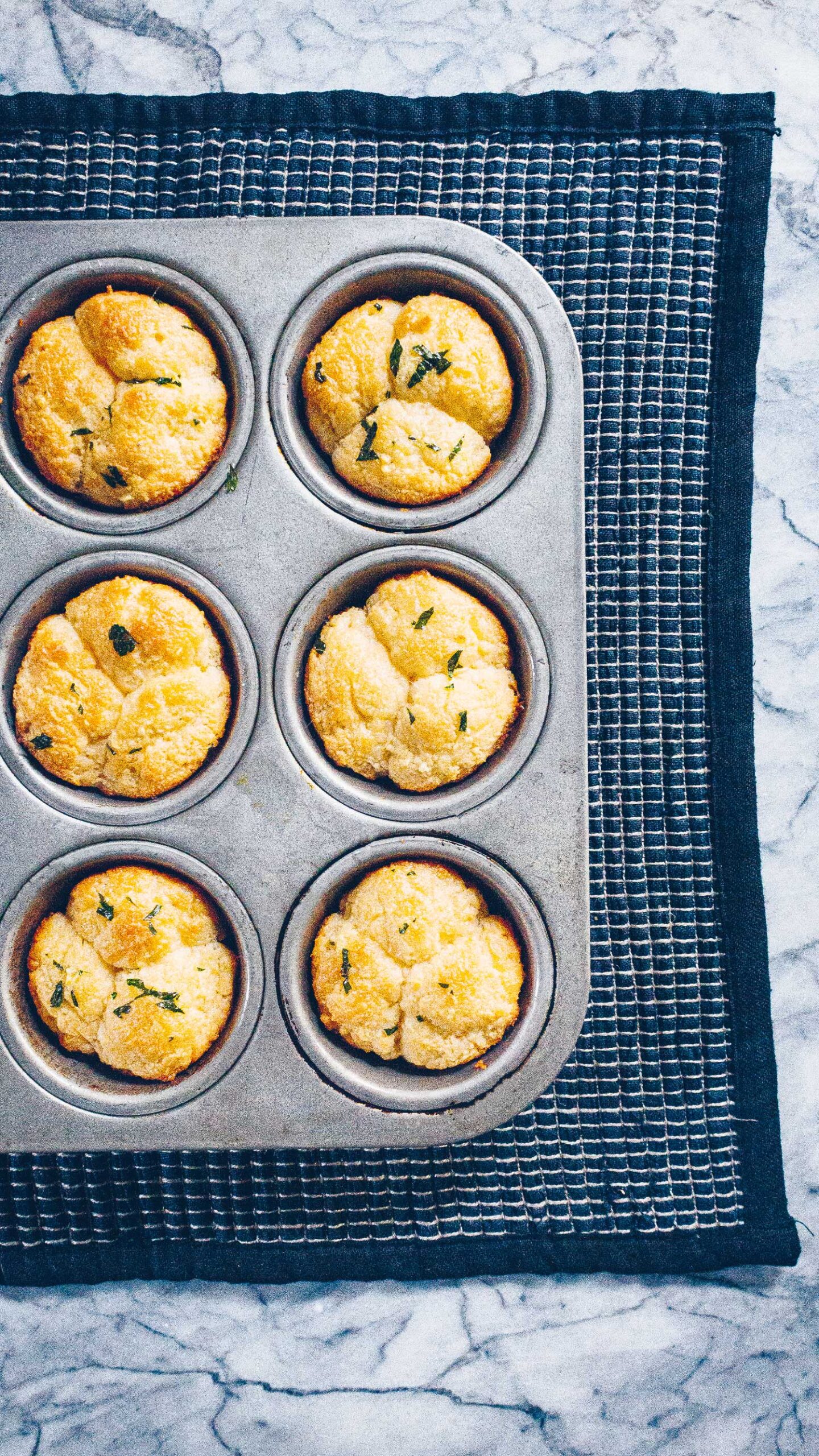 A muffin tin, sitting on a black handwoven placemat on top of a marble countertop, with Cloverleaf rolls fresh from the oven.