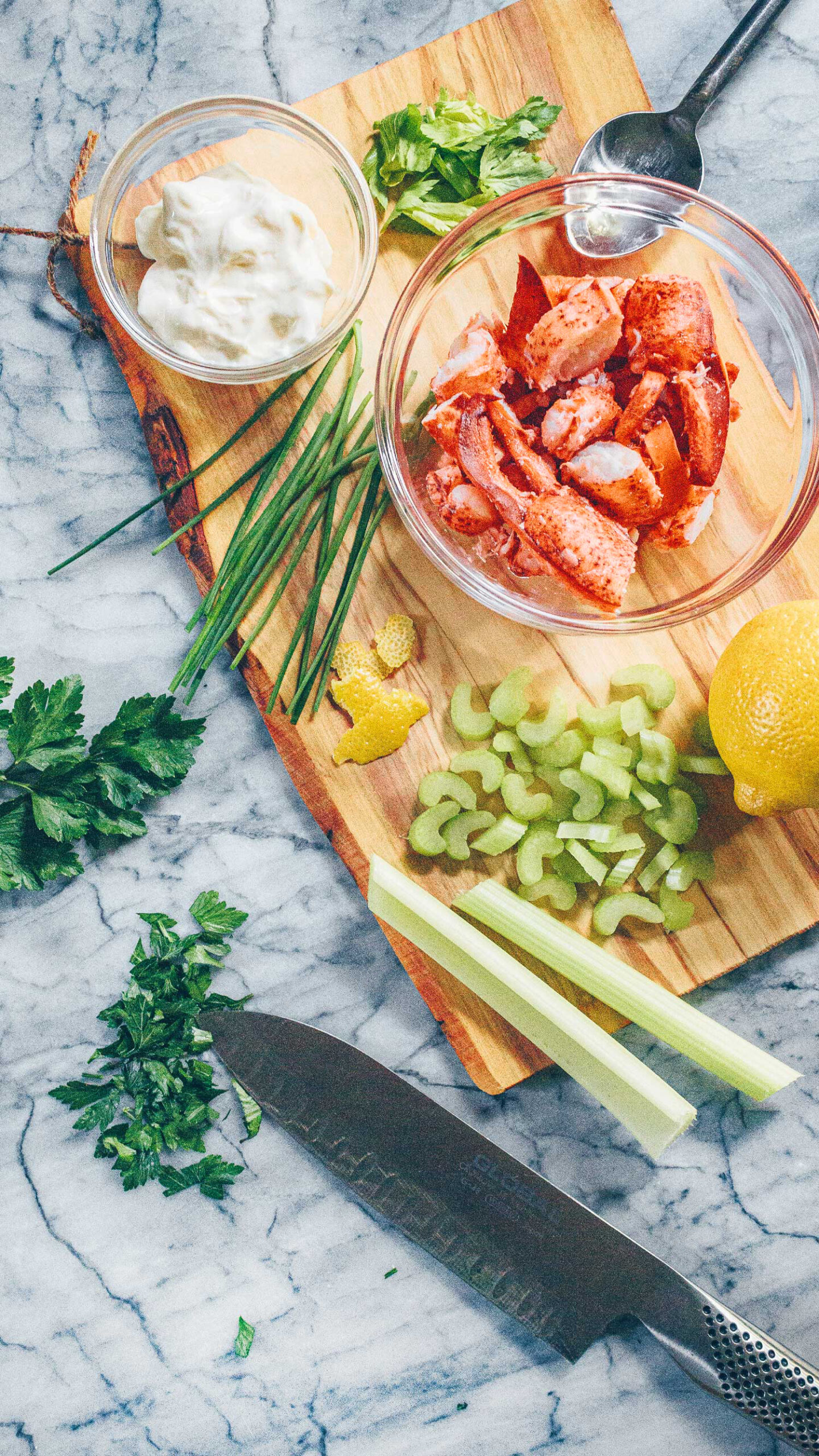 A cutting board with bowls of lobster meat and mayonaisse, surrounded by chives, parsley, lemon, and celery.