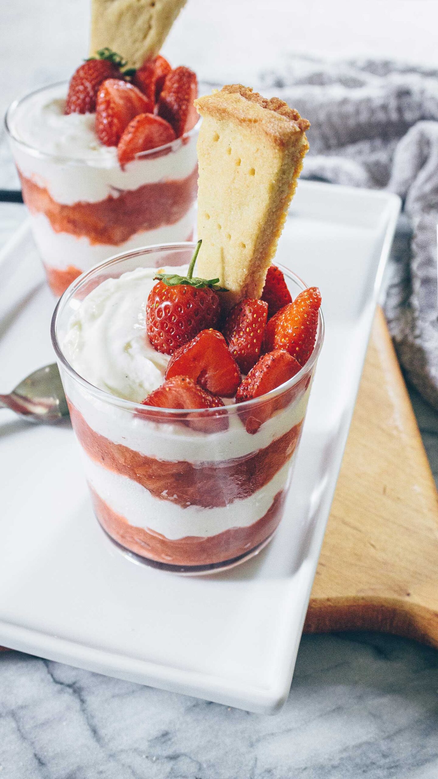A close up of two clear glasses layered with  red Strawberry Rhubarb compote and creamy Cheesecake Cream topped with fresh strawberries.