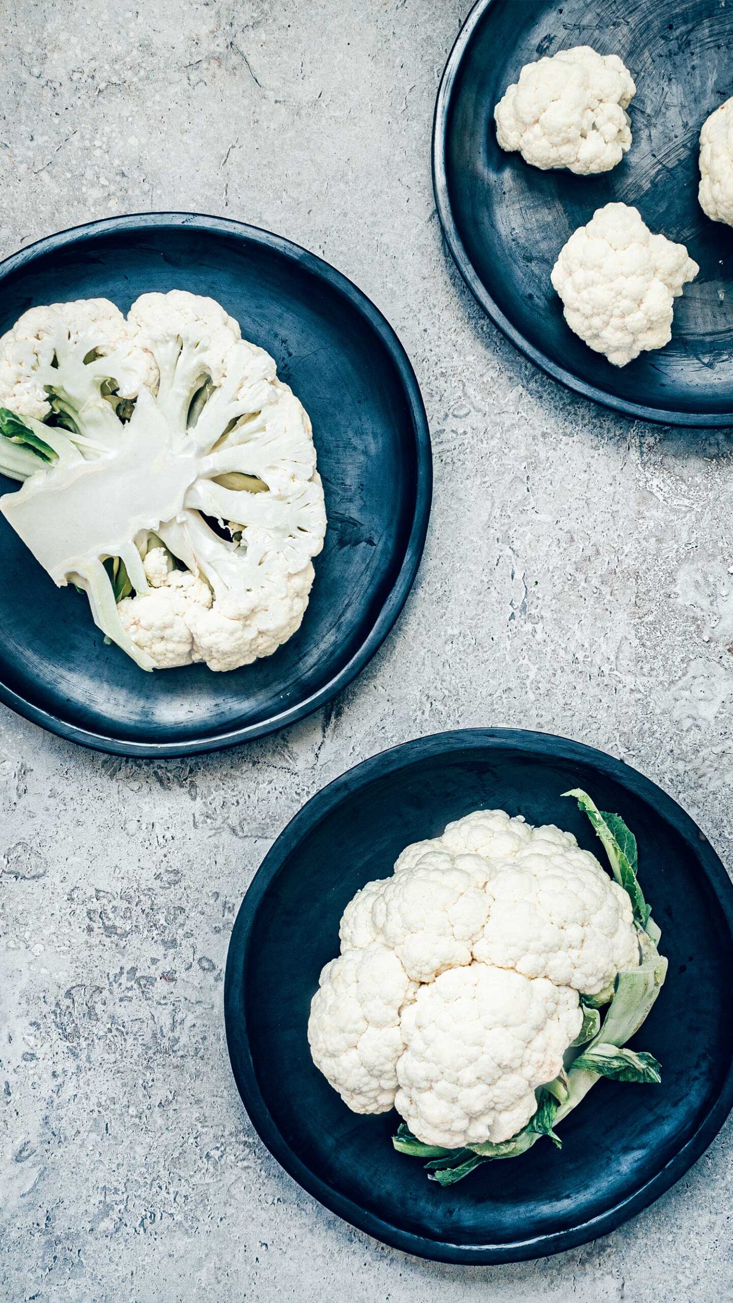 Black pottery plates on a stone background with raw cauliflower decoratively placed. Showing a whole head, individual florets, and a large slice.
