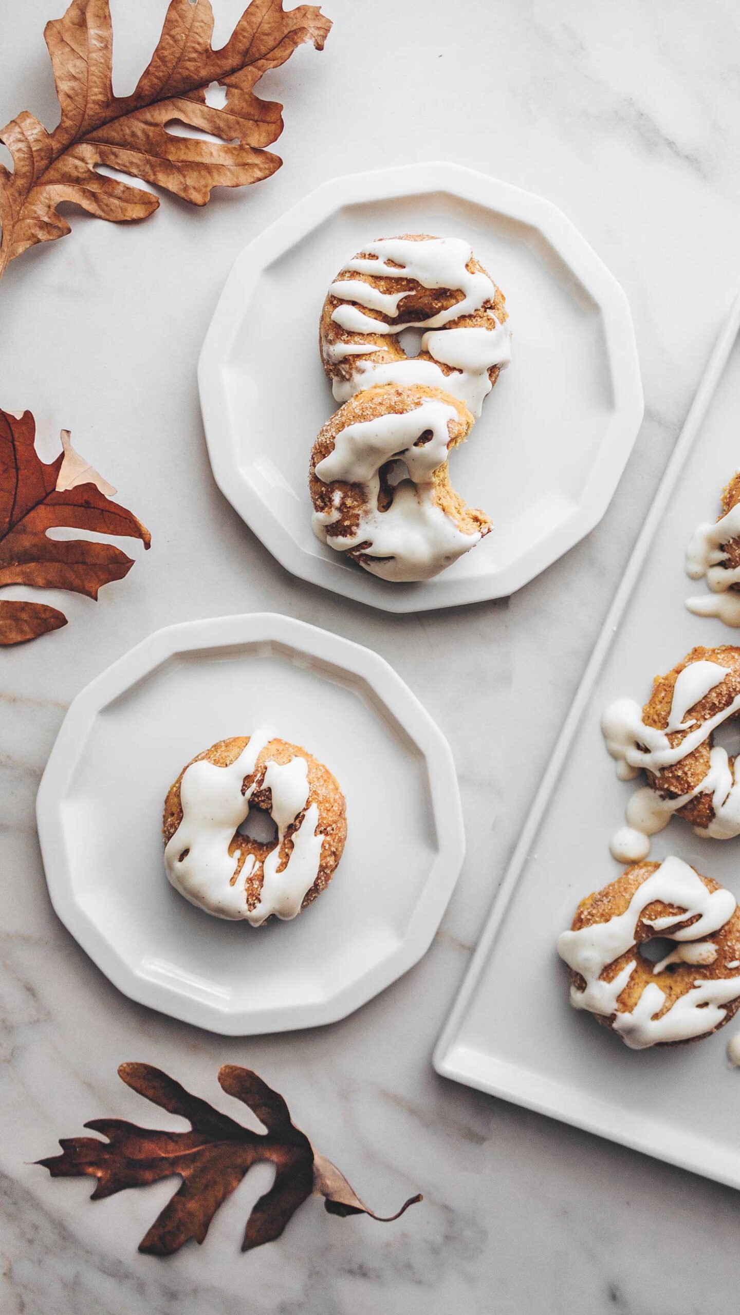 Keto pumpkin spice donuts drizzled with cream cheese glaze on white plates with fall leaves scattered around.