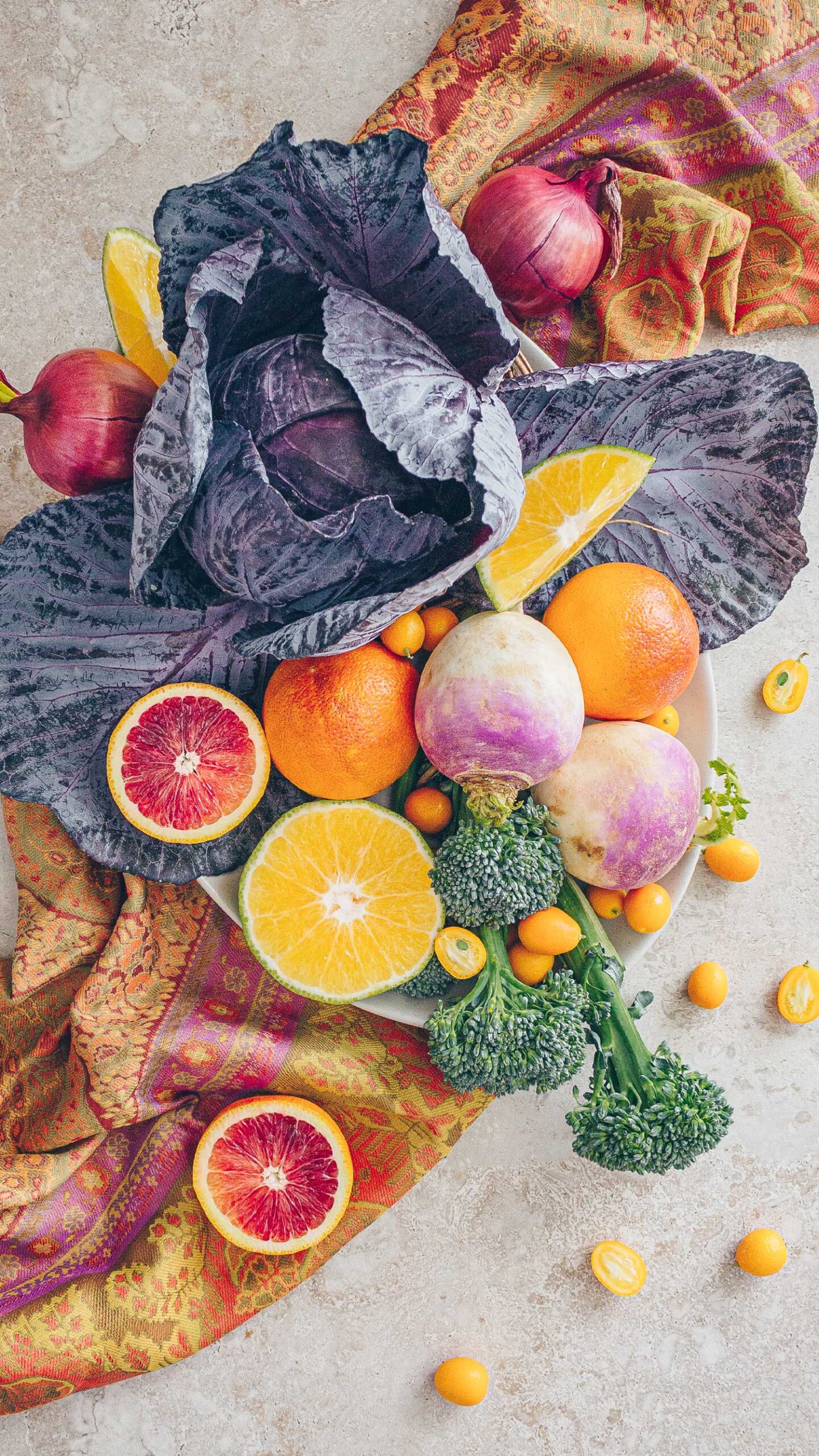 A display of seasonal fruits and vegetables for February: purple cabbage, turnips, broccoli, onions, blood oranges, ugly fruit, and kumquats on a paisley runner.