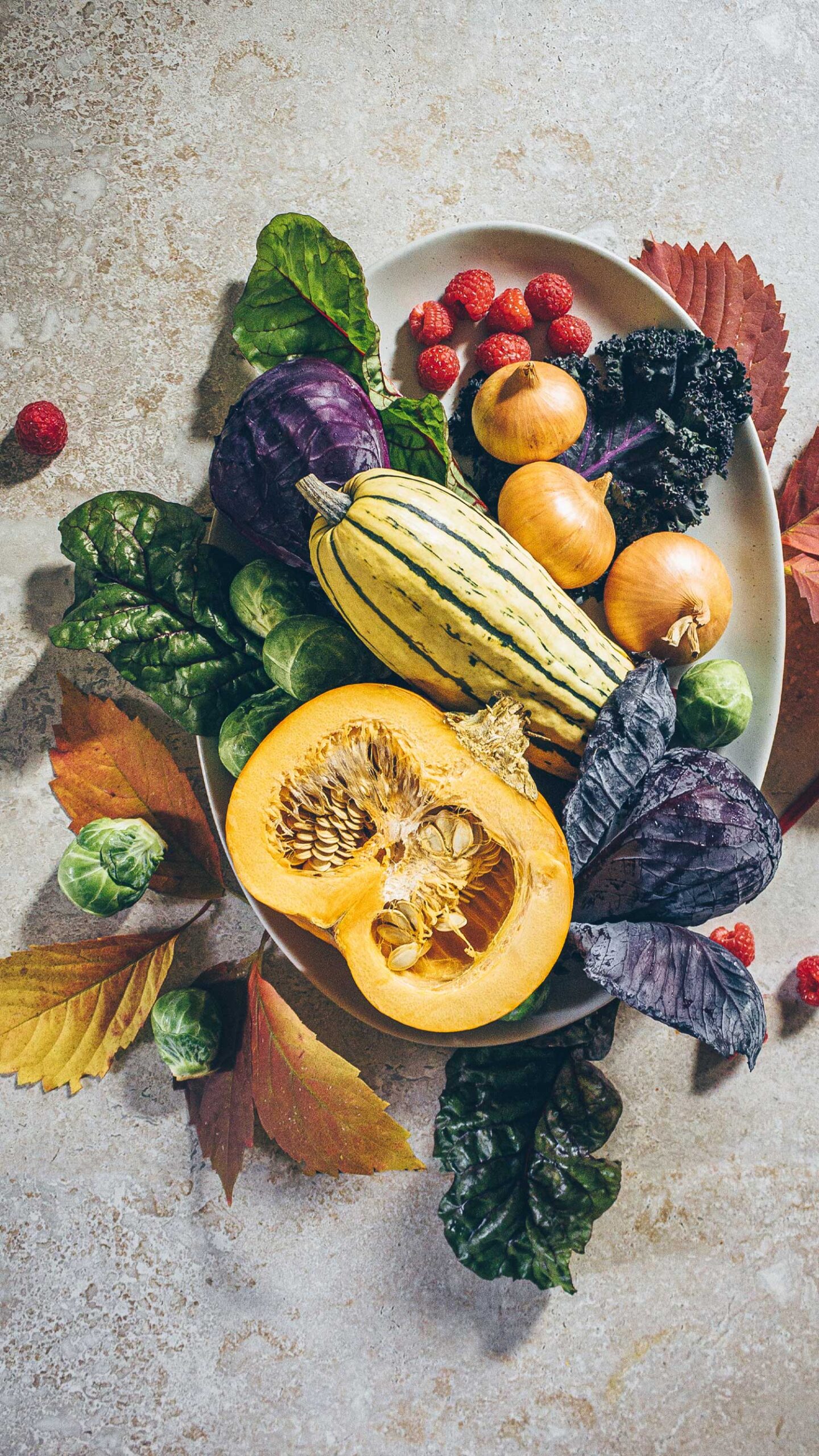 A pottery serving platter with pumpkin, delicate squash, cabbage, onions, kale, raspberries, brussel sprouts, swiss chard, and fall leaves.
