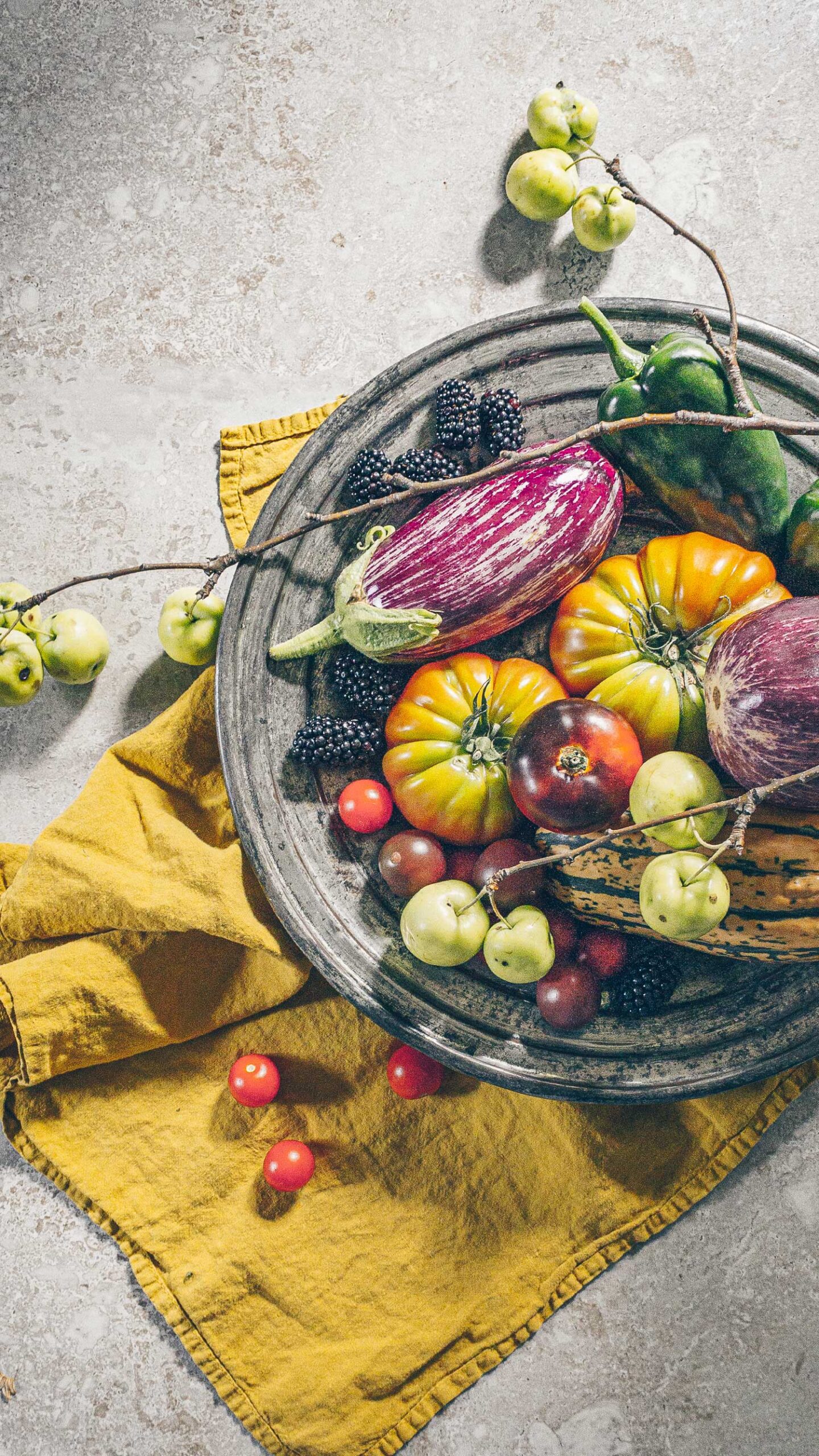 A pewter plate with tomatoes, eggplant, blackberries, wild apples, peppers, and delicata squash, accented by a mustard colored napkin.
