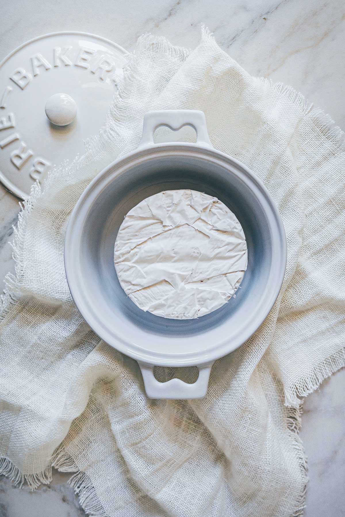A white ceramic brie baker with the top off showing a white, crinkly wheel of Brie cheese inside, waiting to be baked.