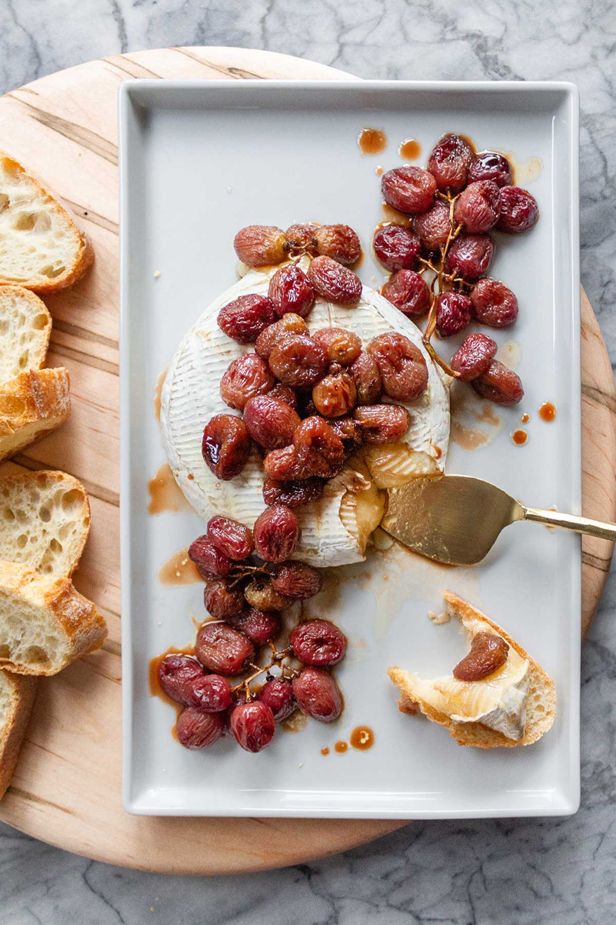 A white ceramic serving plate with a wheel or Baked Brie topped with Roasted Red Grapes. The plate is sitting on top of a round wooden cheese board with slices of fresh baguette.