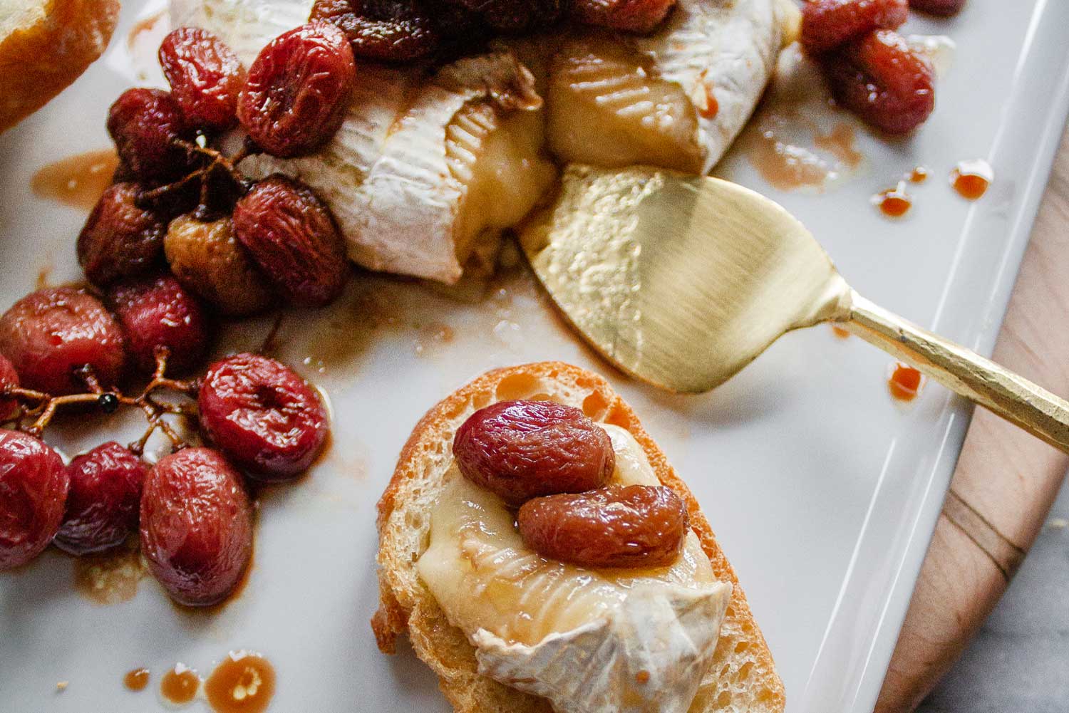 A close up of a slice of baguette topped with a chunk of baked brie cheese and topped with two roasted red grapes.