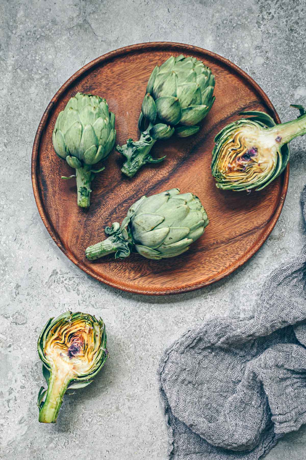 A wooden charger with fresh artichokes on top, two are sliced in half revealing their hearts.