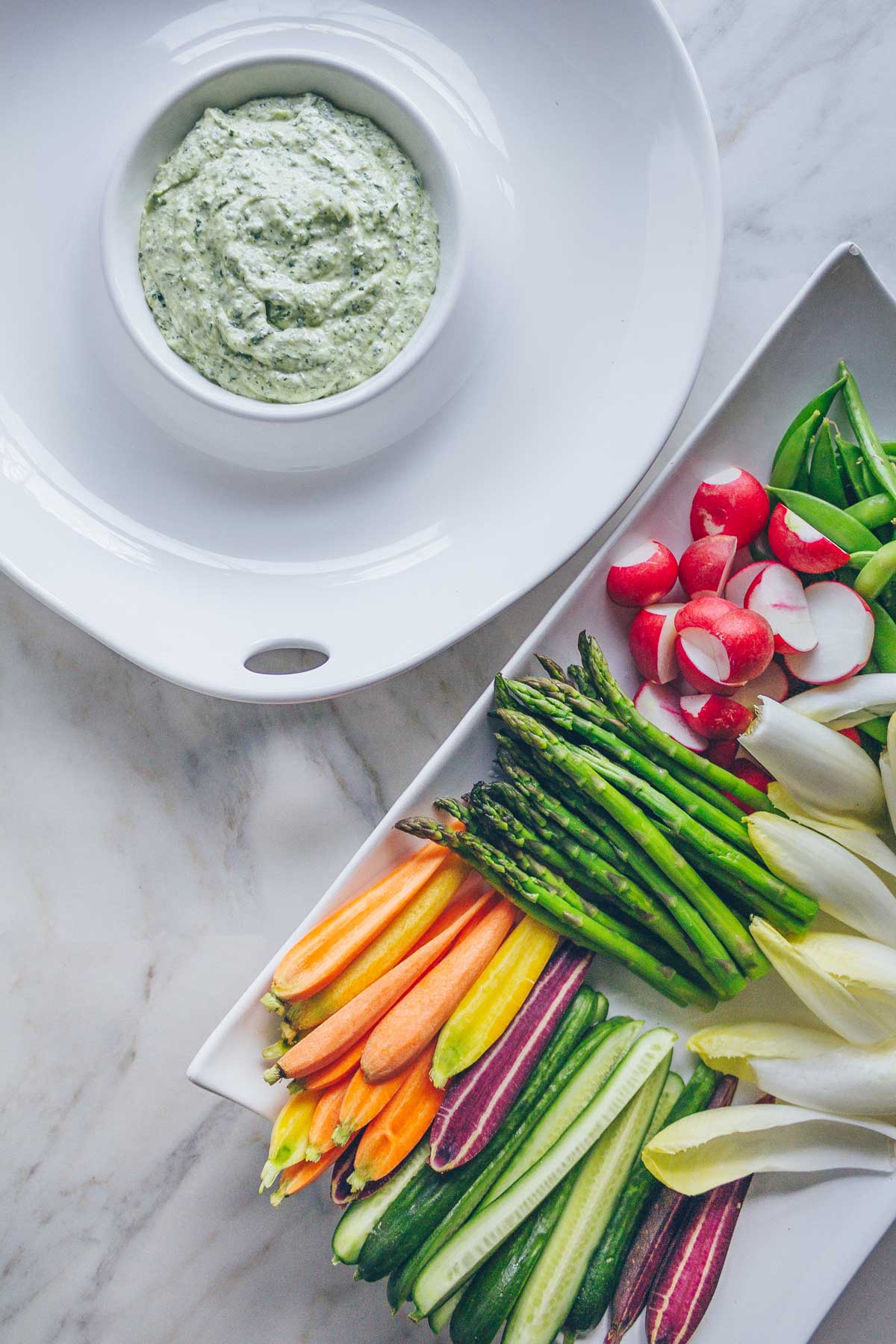 An round white dip platter filled with creamy Greek Goddess Dip, waiting to be filled with vegetables. On 
a platter next to it are heaps of fresh vegetables: multicolor carrots, asparagus, endive, radishes, cucumbers, snap peas, and crispy smashed potatoes.