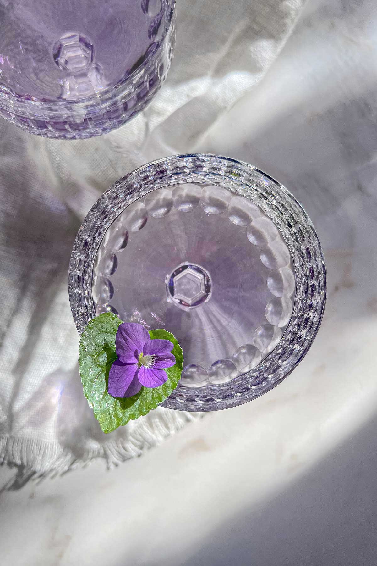 An overhead shot of a lavender hued Aviation Violet Cocktail in a cut crystal coupe glass. A garnish of a violet flower and leaf sit on the side of the glass.