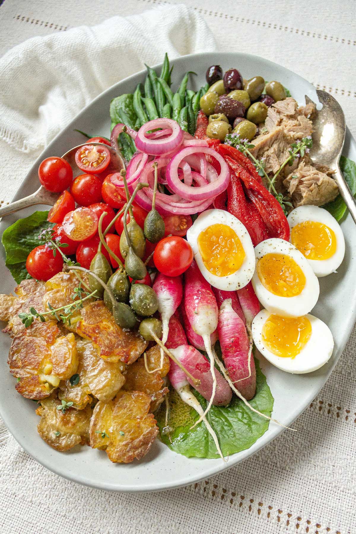 A platter filled with the ingredients for a classic Niçoise salad: green beans, olives, tinned tuna, eggs, potatoes, capers, tomatoes, and garden-fresh greens and herbs.