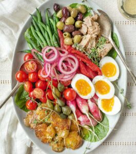 An oval platter with a classic Niçoise salad overflowing with fresh green beans, tomatoes, radishes, crispy smashed potatoes, olives, capers, pickled onions, tuna, and jammy eggs.