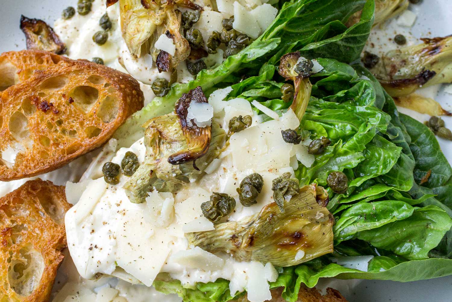 A close up of a romaine lettuce wedge topped with an easy caesar dressing, artichoke hearts, garlic croutons, fried capers, and parmesan cheese. 