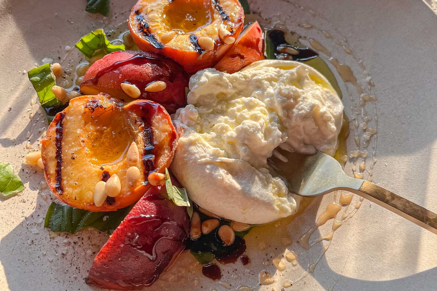 A close up of grilled peach halves and a creamy burrata cheese drizzled with olive oil and honey and scattered with fresh basil and pine nuts.