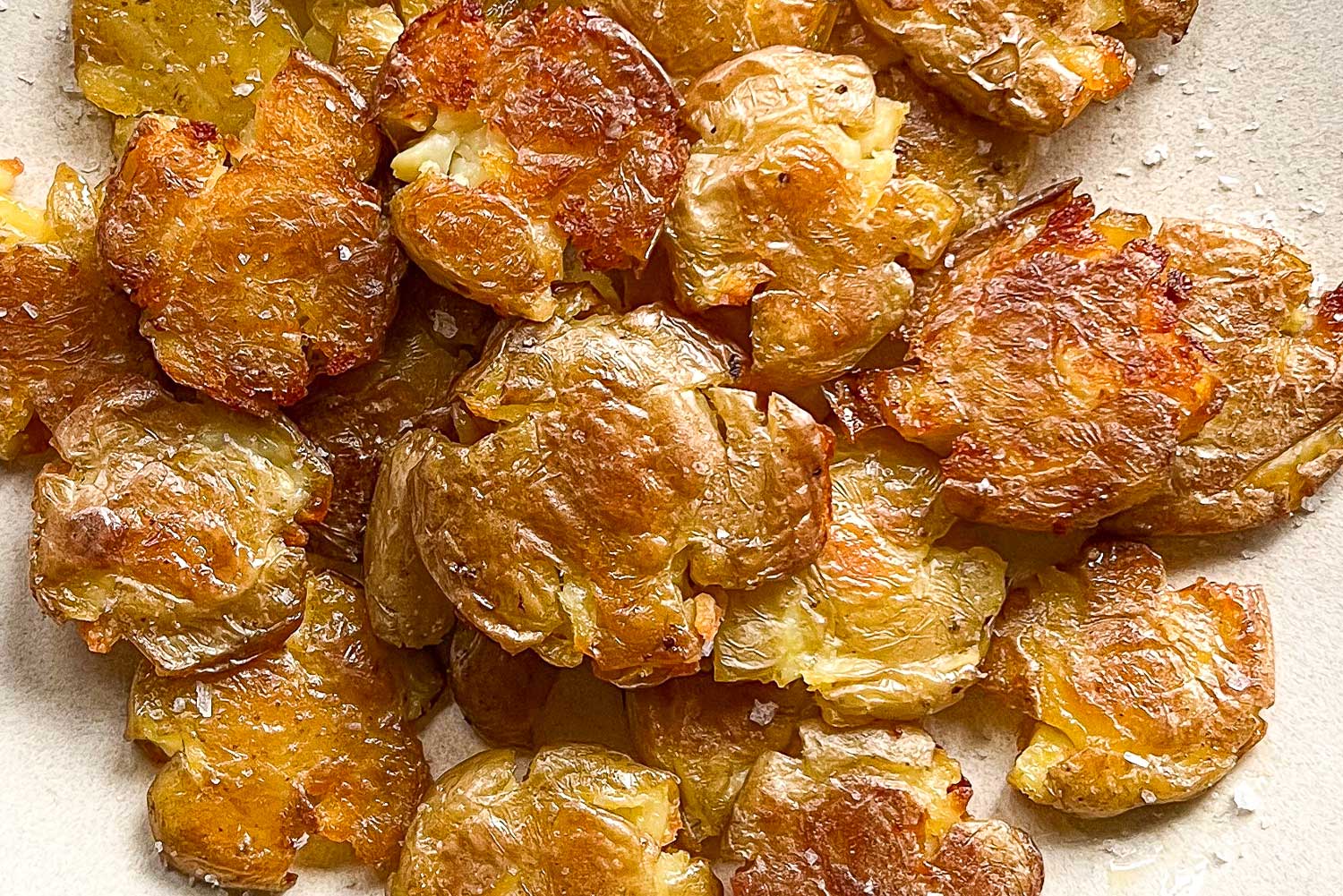 A close up of a pile of roasted crispy smashed potatoes, brown and glistening with olive oil and sprinkled with flakey sea salt.