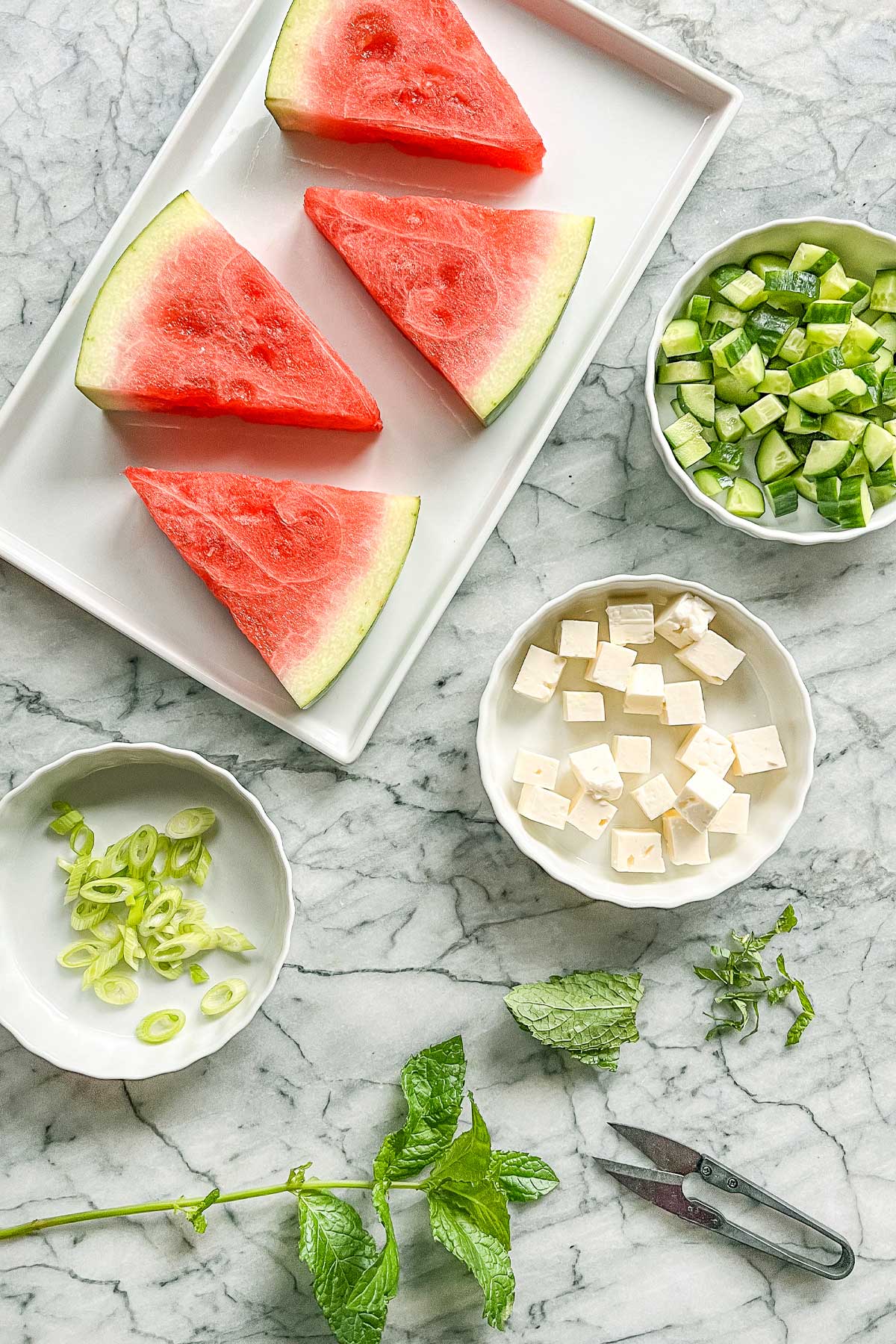 The ingredients for the Watermelon Cucumber Mint salad wedges in white ceramic containers on a marble counter. Watermelon slices, chopped cucumber, feta, sliced scallions, and fresh mint.