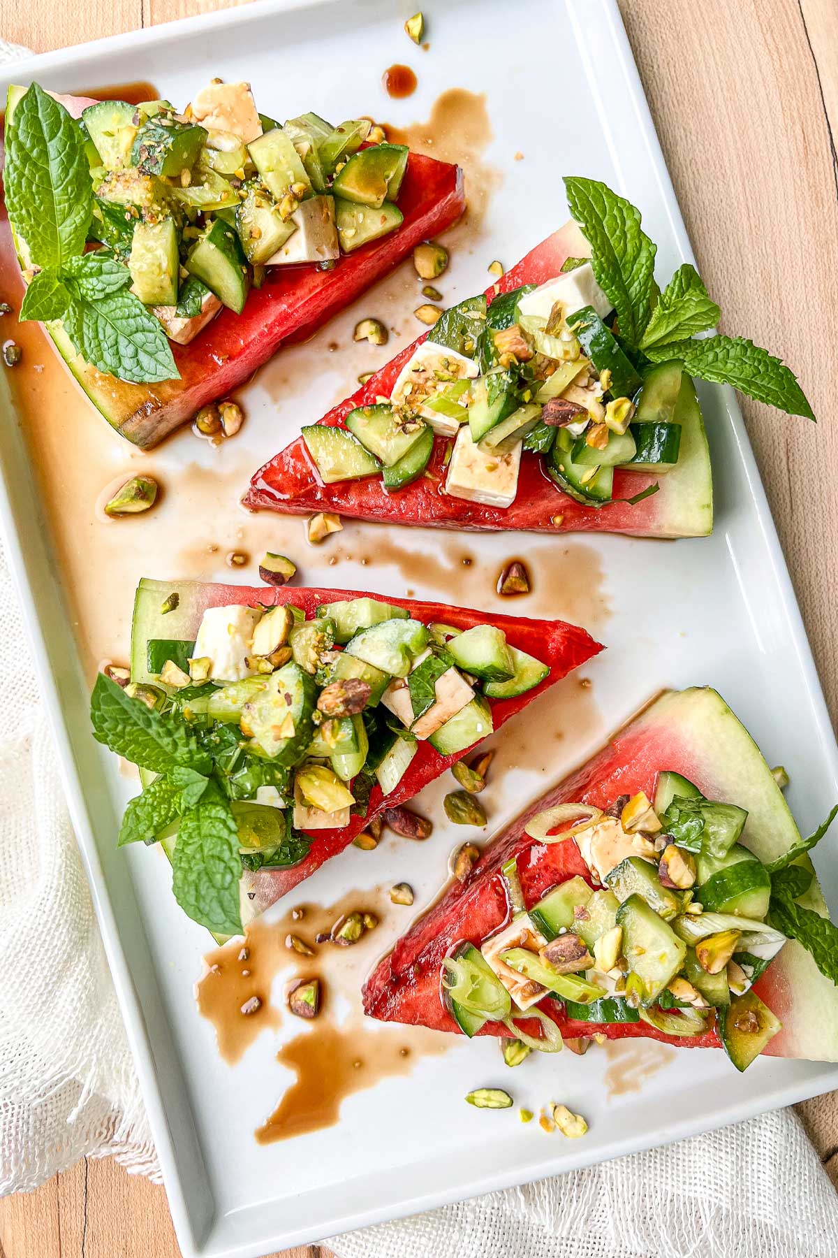 A white rectangular ceramic serving platter with Watermelon Cucumber Mint Salad wedges drizzled in balsamic vinegar and sprinkled with chopped pistachios.