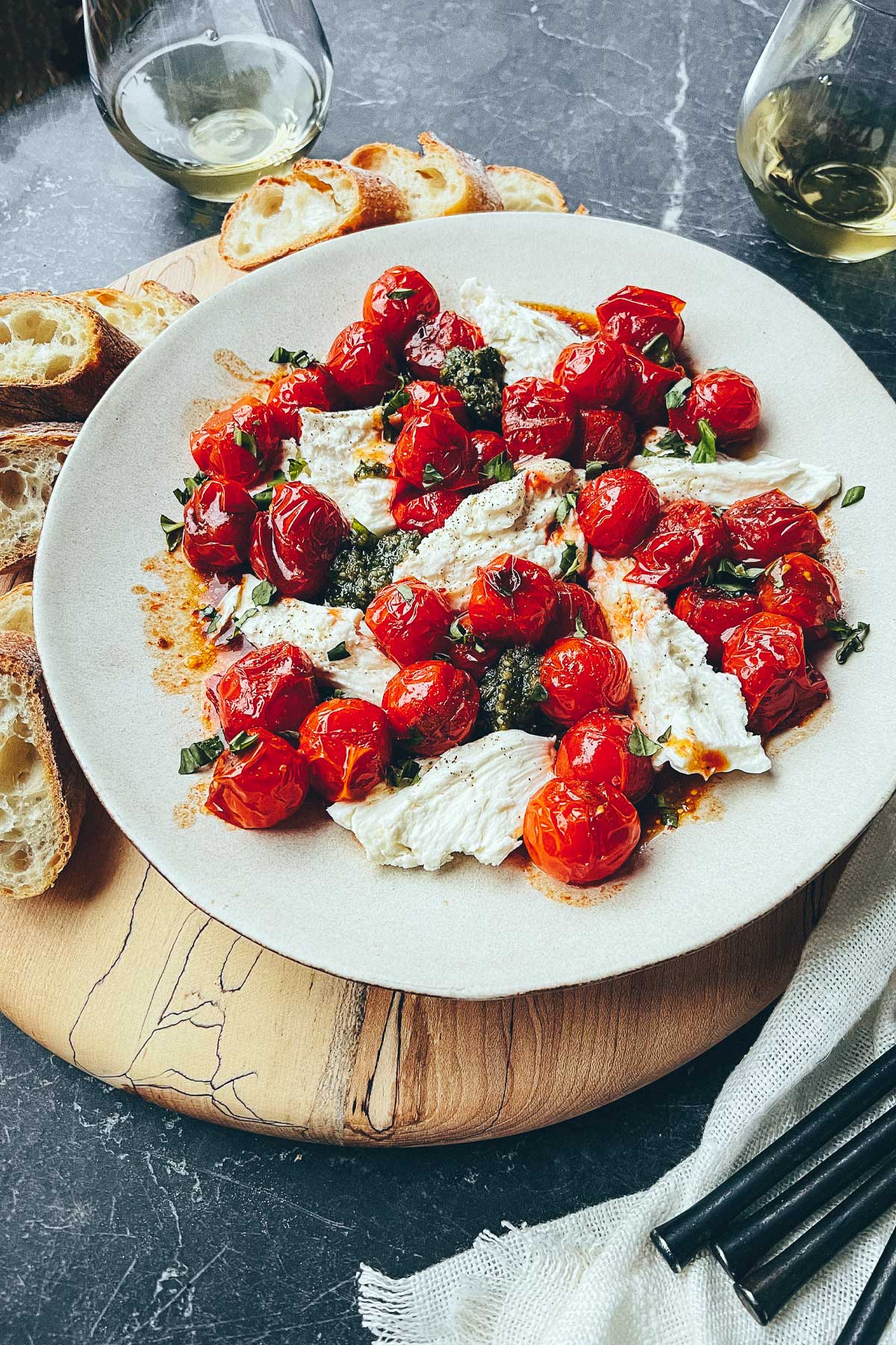 A plate of blistered cherry tomatoes, fresh mozzarella, and pesto sitting on top of a wooden serving board with crusty chunks of fresh baguette.