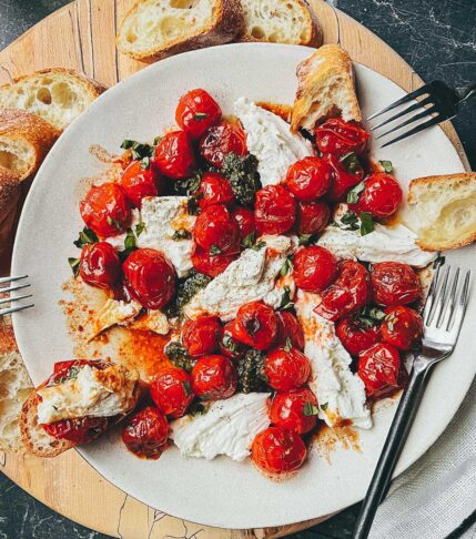 A ceramic plate on a wooden cutting board, layered with blistered red cherry tomatoes, torn pieces of fresh mozzarella cheese, pesto, and fresh basil.