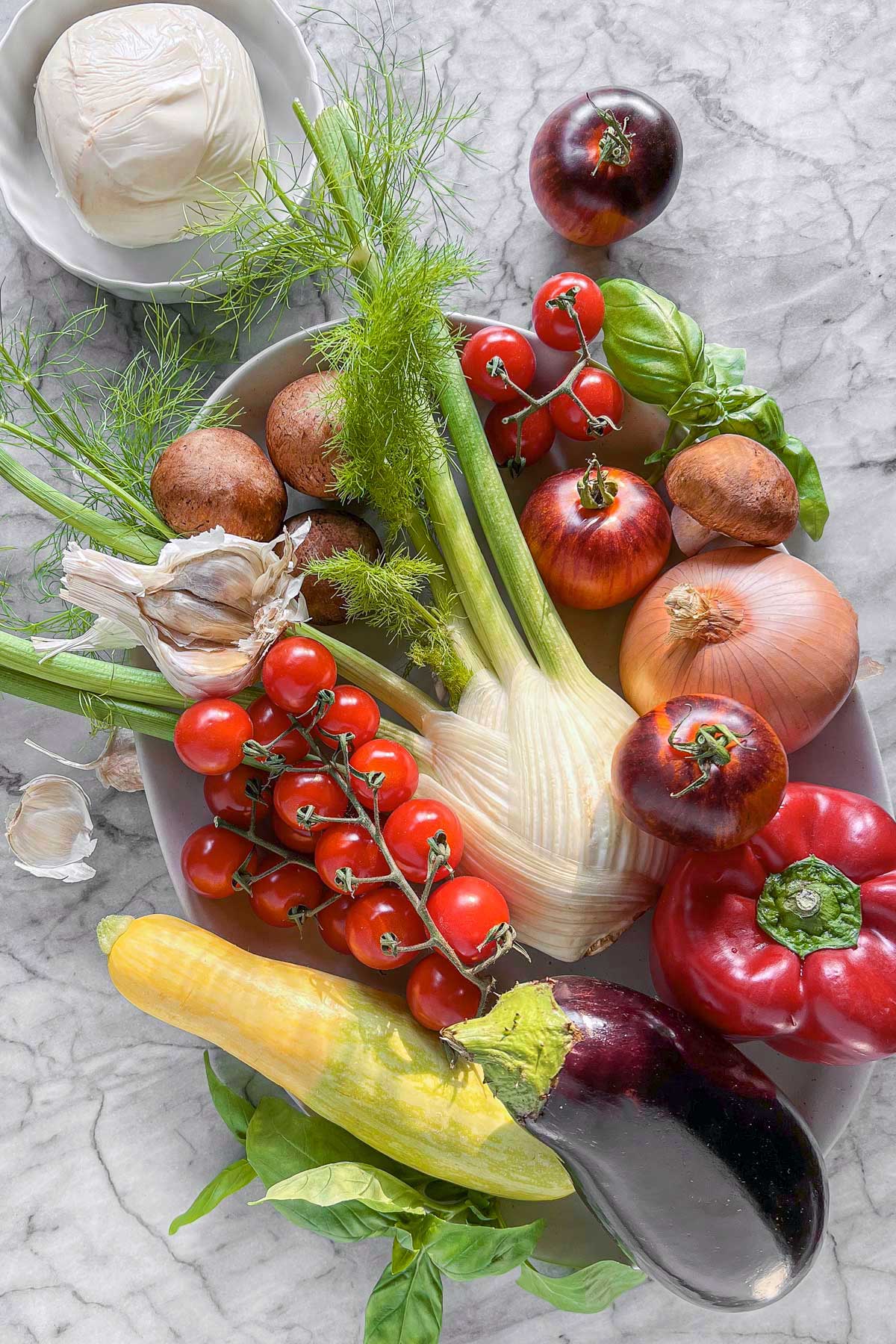 A platter of fresh summer vegetables waiting to be prepped for grilling. Fennel, eggplant, onions, mushrooms, red pepper, and tomatoes sit beside a ball of smoked mozzarella cheese.