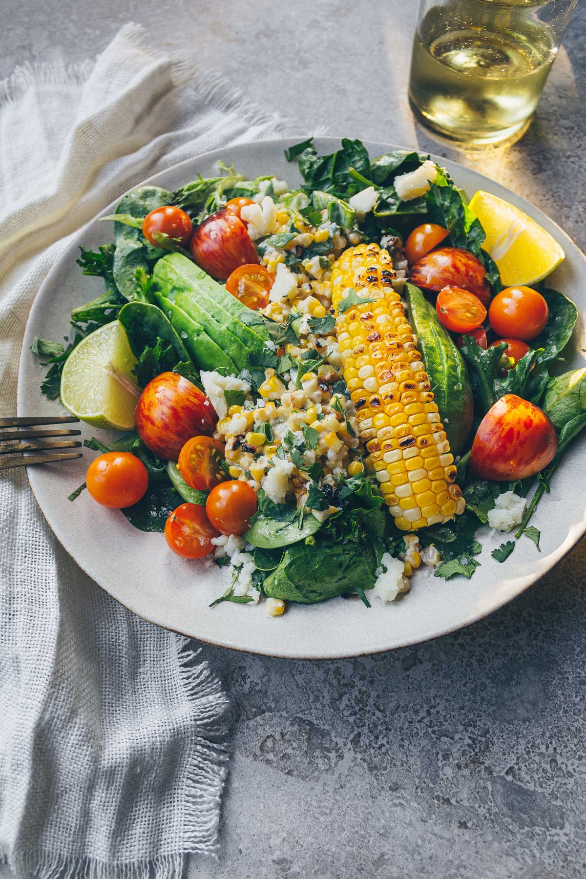 A large plate with a large salad brimming with fresh greens, avocado, grilled corn salad, tomatoes, crumbled cheese, cilantro, and topped with a corn rib.