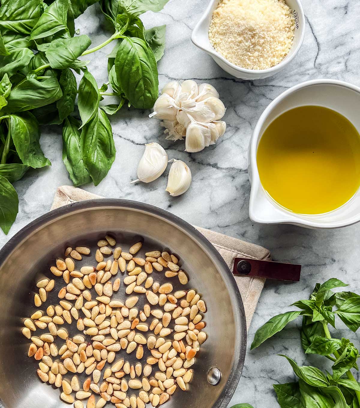 A marble board with fresh basil, pinenuts, garlic, parmesan cheese, and a bowl of extra virgin olive oil.