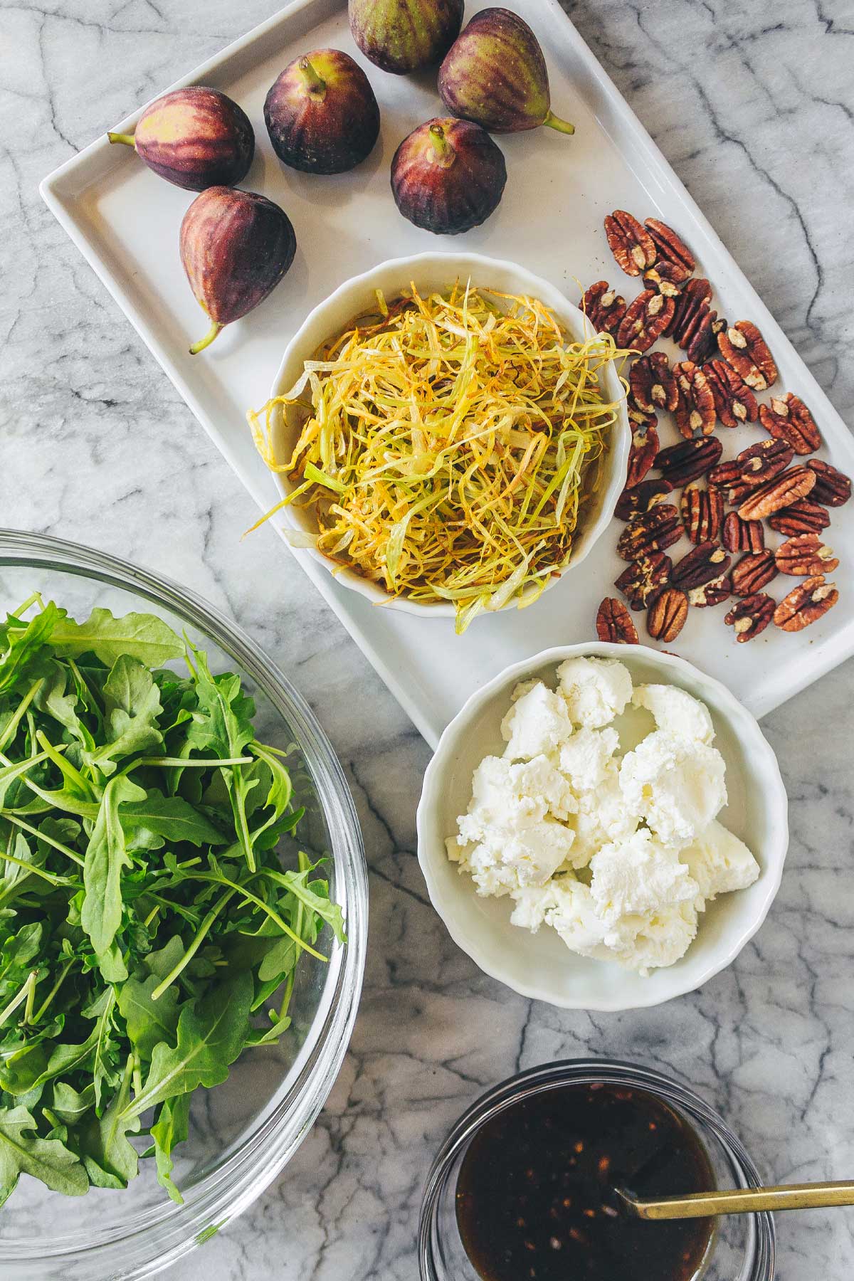 A marble board layered with the ingredients for Fig & Goat Cheese Salad: a bowl of fresh arugula, fresh figs, goat cheese crumbles, toasted pecans, and a bowl of frizzled leeks.