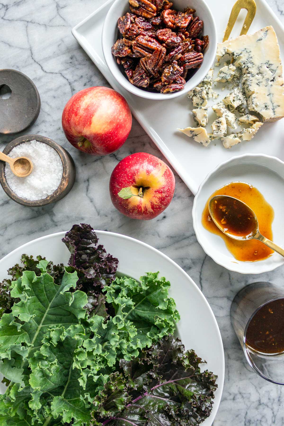 The ingredients for a kale salad with apples laid out on a marble board: fresh kale, apples, crumbled blue cheese, and a bowl of homemade maple pecans.