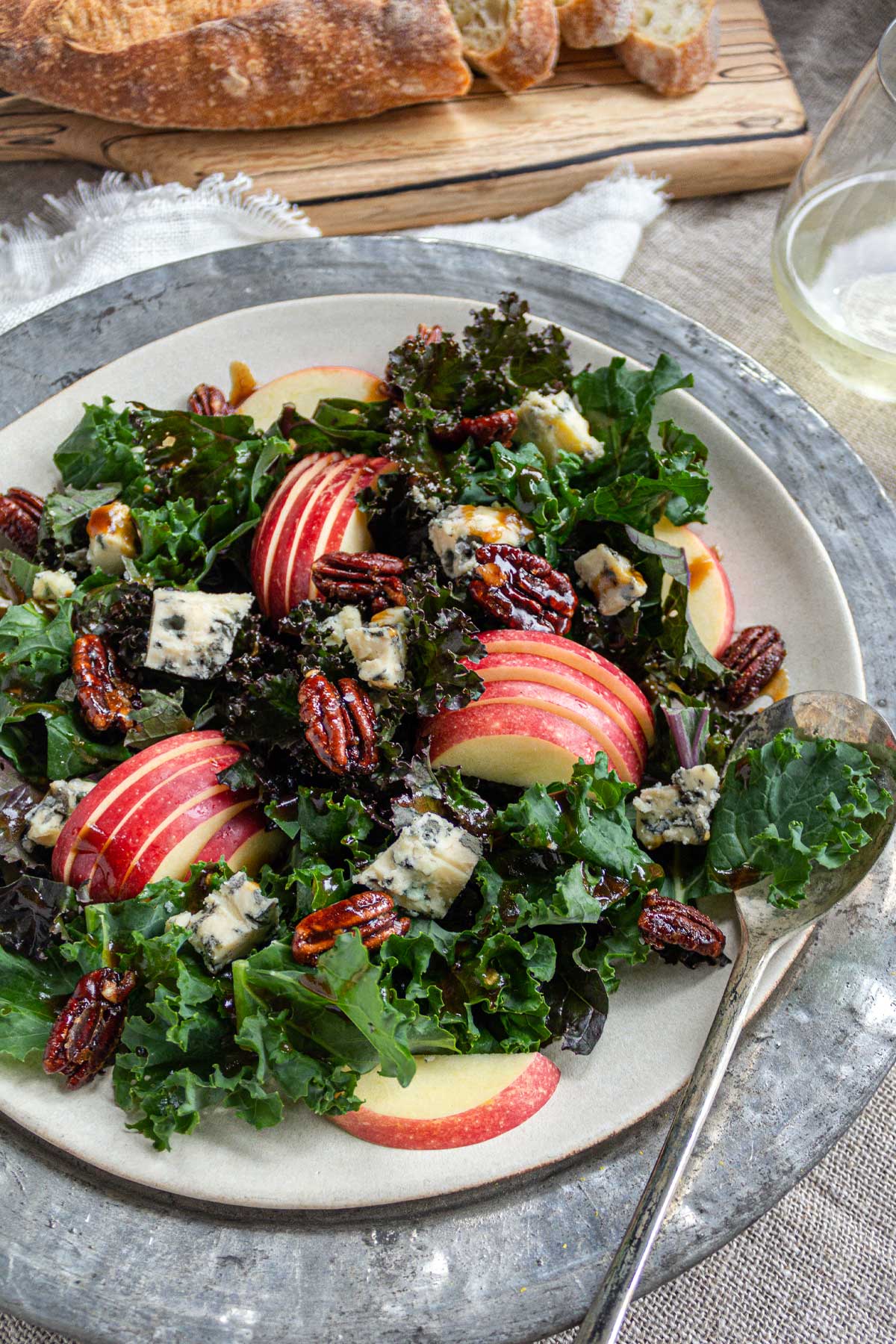 A tablescape with a ceramic plate on a weathered metal charger, filled with a kale salad with apples, blue cheese, and maple pecans.