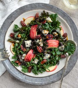A close up of a plate filled with a kale salad with apples, blue cheese crumbles, and candied maple pecans.